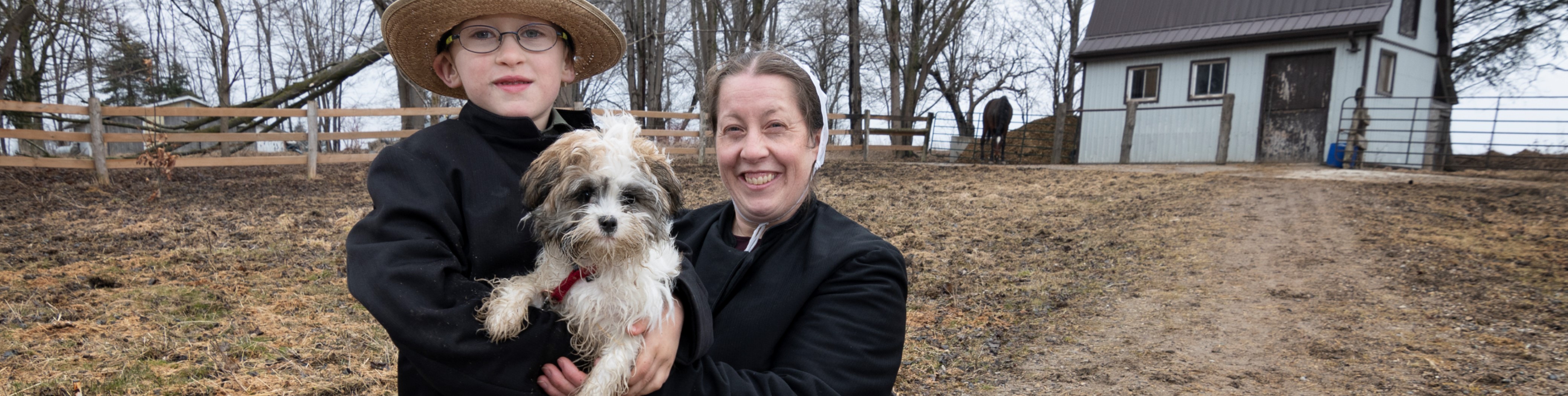 Smiling Mennonite mother and son with dog on lane wane of farm house.