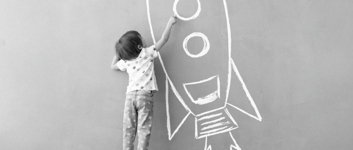 A young boy stands on a box to paint a rocket on the wall.
