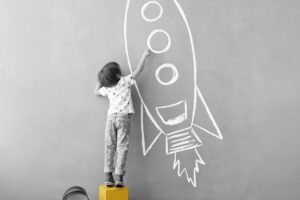 A young boy stands on a box to paint a rocket on the wall.