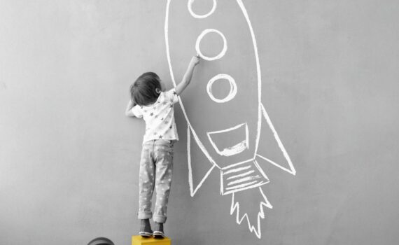 A young boy stands on a box to paint a rocket on the wall.