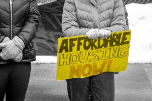 Two protesters stand outside in winter gear. One holds a sign reading “Affordable Housing Now!”