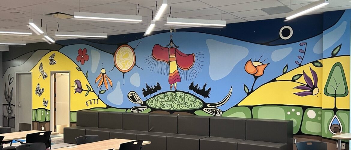 A meeting room with long tables and a long couch by the wall. The wall has a colourful Indigenous mural with flowers, a woman dancing on a turtle’s back, and butterflies.