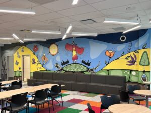 A meeting room with long tables and a long couch by the wall. The wall has a colourful Indigenous mural with flowers, a woman dancing on a turtle’s back, and butterflies.
