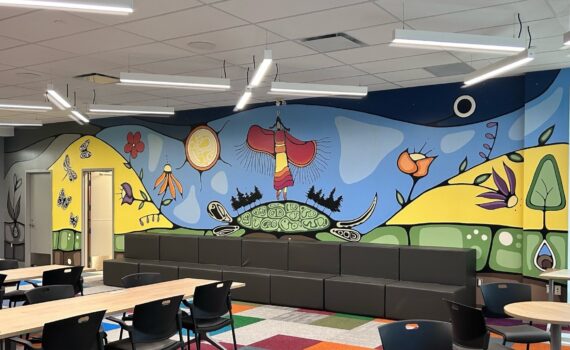 A meeting room with long tables and a long couch by the wall. The wall has a colourful Indigenous mural with flowers, a woman dancing on a turtle’s back, and butterflies.