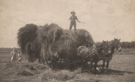A vintage, sepia photograph of a farmer standing on a cart with a huge pile of hay.